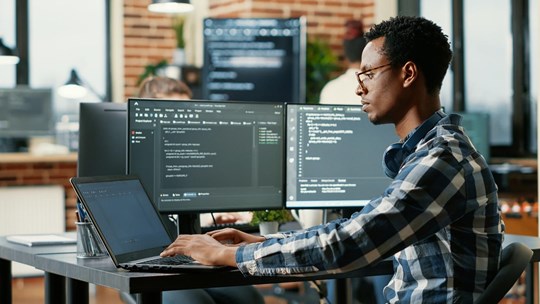 Portrait of african american developer using laptop to write code sitting at desk with multiple screens parsing algorithm in software agency. Coder working on user interface using portable computer.; Shutterstock ID 2108122673; purchase_order: N/A; job: TE-B; client: Tietoevry; other: 