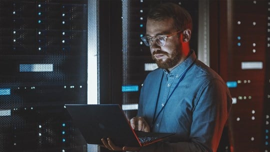 Bearded IT Specialist in Glasses is Working on Laptop in Data Center while Standing Near Server Rack. Running Diagnostics, Doing Maintenance Work. Emergency Red Light from Side Illuminating Specialist